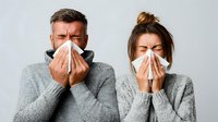 Couple in gray sweaters blowing noses into tissues simultaneously, showing symptoms of seasonal cold or flu against light background.