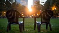 Two empty plastic chairs on green grass in urban park at sunset with city skyline and glowing lights in background.