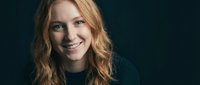 Young blonde woman with curly hair smiling warmly at camera against dark background. Professional headshot portrait for business and lifestyle content.