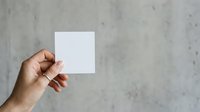 A person with medium skin tone holds a blank white square card against a textured concrete wall background, suitable for branding, mockups, and promotional material presentations.