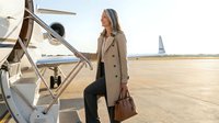 Mature woman in beige coat standing on airplane stairs at airport tarmac, holding brown leather bag, ready for travel or business trip.