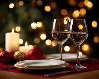 Romantic holiday table setting with wine glasses, white plates, and festive decorations against blurred Christmas lights and burning candles on burgundy tablecloth.