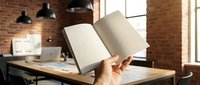 Person holding open blank notebook in modern loft office with brick walls, wooden desk, computer and pendant lights, suitable for business, education and creative workspace concepts.