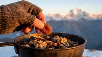 Gloved hand stirring roasted nuts and chickpeas in cast iron skillet at mountain summit during golden hour sunrise, outdoor camping cooking.