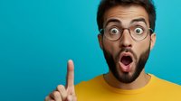 Young surprised Caucasian man with glasses pointing up with index finger showing shocked facial expression on turquoise background for advertising concepts.