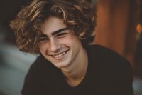 Young handsome man with curly hair smiling warmly in cozy indoor setting. Genuine happiness and positive emotions captured in natural portrait lighting.