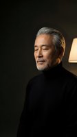 Mature Asian businessman with gray beard in black turtleneck looking contemplative in warm indoor lighting for corporate and lifestyle content.