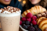 Creamy coffee drink with cocoa powder and whipped cream served alongside fresh croissants, raspberries and blackberries on elegant breakfast table setting.
