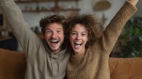 Young couple celebrating with raised arms and joyful expressions in cozy living room setting. Happy man and woman showing excitement and success.