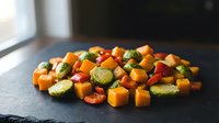 Colorful roasted vegetables including Brussels sprouts, sweet potato, cherry tomatoes and carrots arranged on dark slate surface for healthy cooking.
