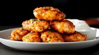 Crispy golden chicken nuggets garnished with fresh herbs, stacked on white plate against dark background. Close-up food photography shows juicy texture and caramelized coating.