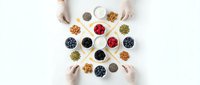 Hands reaching for colorful superfood bowls with berries, nuts and seeds arranged in geometric pattern on white background for healthy eating concepts.