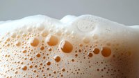 Frothy beer foam with bubbles in macro photography showing detailed texture and natural amber color against light background. Fresh draft beer pour.