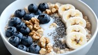 Fresh blueberries, sliced banana, walnuts and chia seeds on creamy oatmeal porridge in white ceramic bowl, healthy breakfast meal closeup.