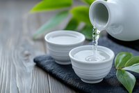 Traditional tea ceremony with white ceramic teapot pouring hot water into cups on dark slate board with fresh green leaves on wooden table background.