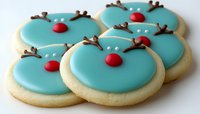 Festive sugar cookies decorated with turquoise icing, brown antlers and red nose design creating playful reindeer faces, arranged on white surface for holiday baking.