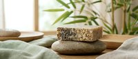 Two natural soap bars stacked on wooden surface with green plant and soft fabric, spa and wellness product for skincare routines.