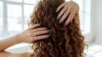 Young woman with beautiful curly brown hair touching her locks in bright bathroom. Hair care, beauty routine and styling concept for salon advertising.