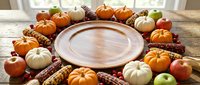 Autumn harvest arrangement with empty wooden plate surrounded by pumpkins, gourds, corn, and apples on rustic table for fall seasonal dining and Thanksgiving table settings.