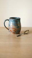Handmade ceramic mug with blue and orange glazed finish sits on wooden table beside silver spoon against neutral background for kitchen decor.