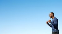 Athletic male runner with dark skin tone jogging outdoors against clear blue sky, wearing casual athletic wear for fitness, exercise, and wellness content.
