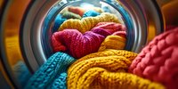 Colorful knitted wool sweaters and clothing inside washing machine drum. Close up view through round door glass shows vibrant textiles in laundry cycle.