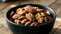 Mixed nuts including walnuts and pecans in dark ceramic bowl on rustic wooden table surface for healthy snacking and cooking ingredients.