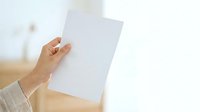 Person holding blank white paper sheet in hand against blurred interior background, suitable for mockup, branding, and presentation designs.