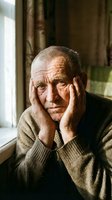Elderly Caucasian man with hands on face showing worry, stress or depression sitting by window in warm indoor lighting for mental health concepts.