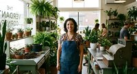 Young Hispanic woman plant shop owner in denim apron standing proudly in green nursery filled with potted plants and hanging ferns for small business concept.