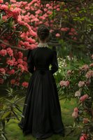 Woman in Victorian black dress stands among blooming pink flowers in garden, view from behind. Dark romantic atmosphere with lush spring blossoms surrounds figure.