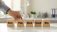 Business professional arranging wooden blocks spelling Start Plan Work Check Win strategy concept for project management and goal achievement success.