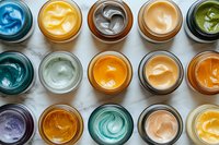 Colorful collection of cosmetic face masks and beauty products in glass jars arranged in rows on marble surface, showing various textures and swirls.
