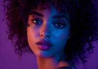 Young African American woman with natural curly hair in dramatic purple and blue neon lighting, showcasing vibrant makeup and expressive eyes for beauty and fashion concepts.