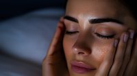 Young Middle Eastern woman with closed eyes and glowing skin touching face in dark moody lighting, showcasing natural beauty and skincare concept.