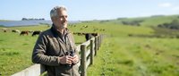 Mature Caucasian farmer standing by wooden fence in green pasture with grazing cattle, rural countryside landscape, agricultural lifestyle and livestock farming.