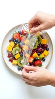 Hands wrapping fresh fruit platter with plastic film, featuring strawberries, blueberries, kiwi and mango on ceramic plate for food storage and preservation.