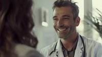 Smiling mature male doctor with gray hair and beard wearing white coat and stethoscope consulting with patient during medical appointment in modern clinic office.