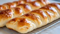 Freshly baked soft bread rolls with golden brown glossy crust arranged in row on cooling rack, close up view showing texture and shine of homemade pastry.