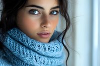 Young woman with natural makeup wearing cozy blue knitted scarf looking at camera with gentle expression, dark hair and blue eyes in soft window light.