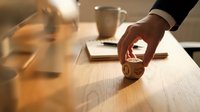 Professional businessman hand placing golden coin on wooden desk in warm office lighting with blurred background creating investment and savings concept.