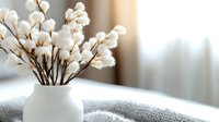 Delicate cotton stems in white ceramic vase on gray knitted texture near window with soft natural light, creating cozy minimalist home atmosphere.