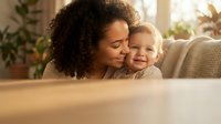 Young African American mother with curly hair embracing happy blonde toddler on cozy living room sofa. Warm family bonding moment at home.