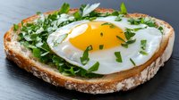 Sunny side up egg on rustic bread slice garnished with fresh herbs and microgreens, served on dark surface, close up view of breakfast or brunch dish.