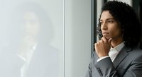 Thoughtful businesswoman with curly dark hair and medium skin tone wearing gray blazer, hand near chin in contemplative pose by window, reflecting on work decisions.