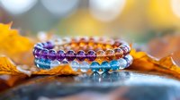 Colorful crystal healing bracelets with purple, amber and blue beads arranged on autumn maple leaves with soft bokeh background, spiritual wellness concept.