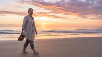 Middle-aged Caucasian man in casual linen shirt walking barefoot on sandy beach at golden hour sunset, holding shoes in hand, ocean waves in background.