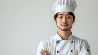 Young Asian male chef in traditional white uniform and toque standing confidently with arms crossed against neutral background, professional culinary portrait.