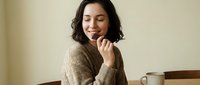 Young Asian woman in cozy beige sweater enjoying peaceful moment with eyes closed, hand near chin in contemplative pose. Coffee cup on wooden table creates warm atmosphere.