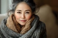 Mature Asian woman wearing cozy knit scarf and sweater smiling warmly at camera, natural beauty portrait showcasing graceful aging and authentic expression.